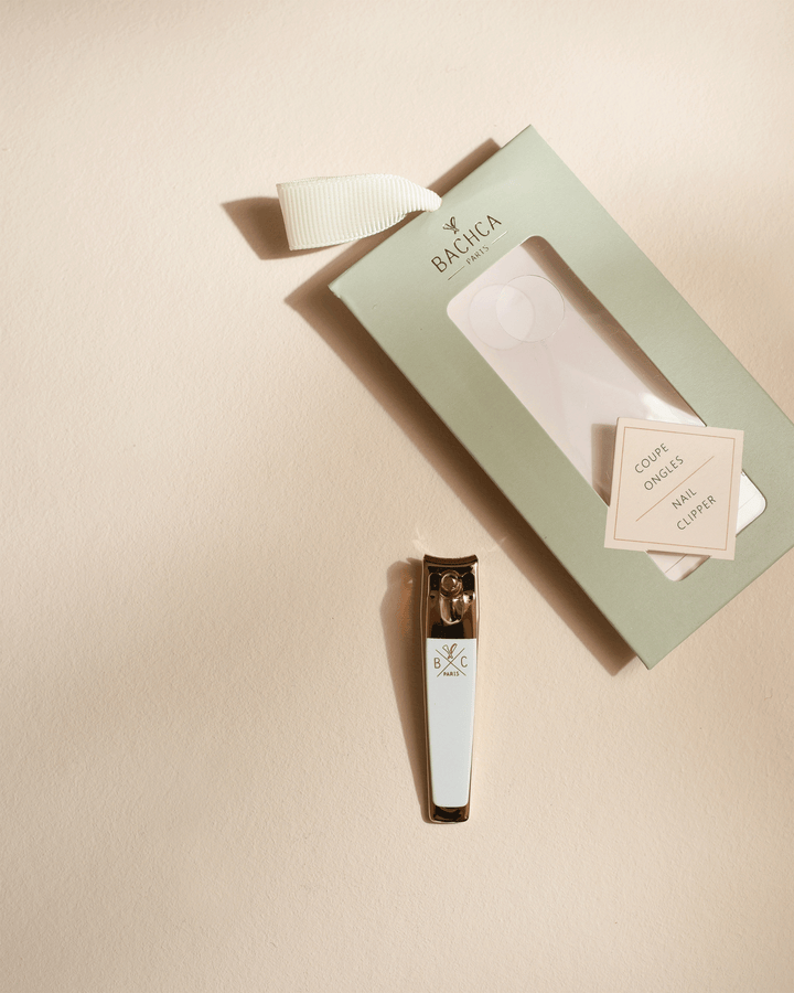 Gold and white nail clipper displayed next to its empty sage green box on a neutral, warm beige surface with elegant shadows.
