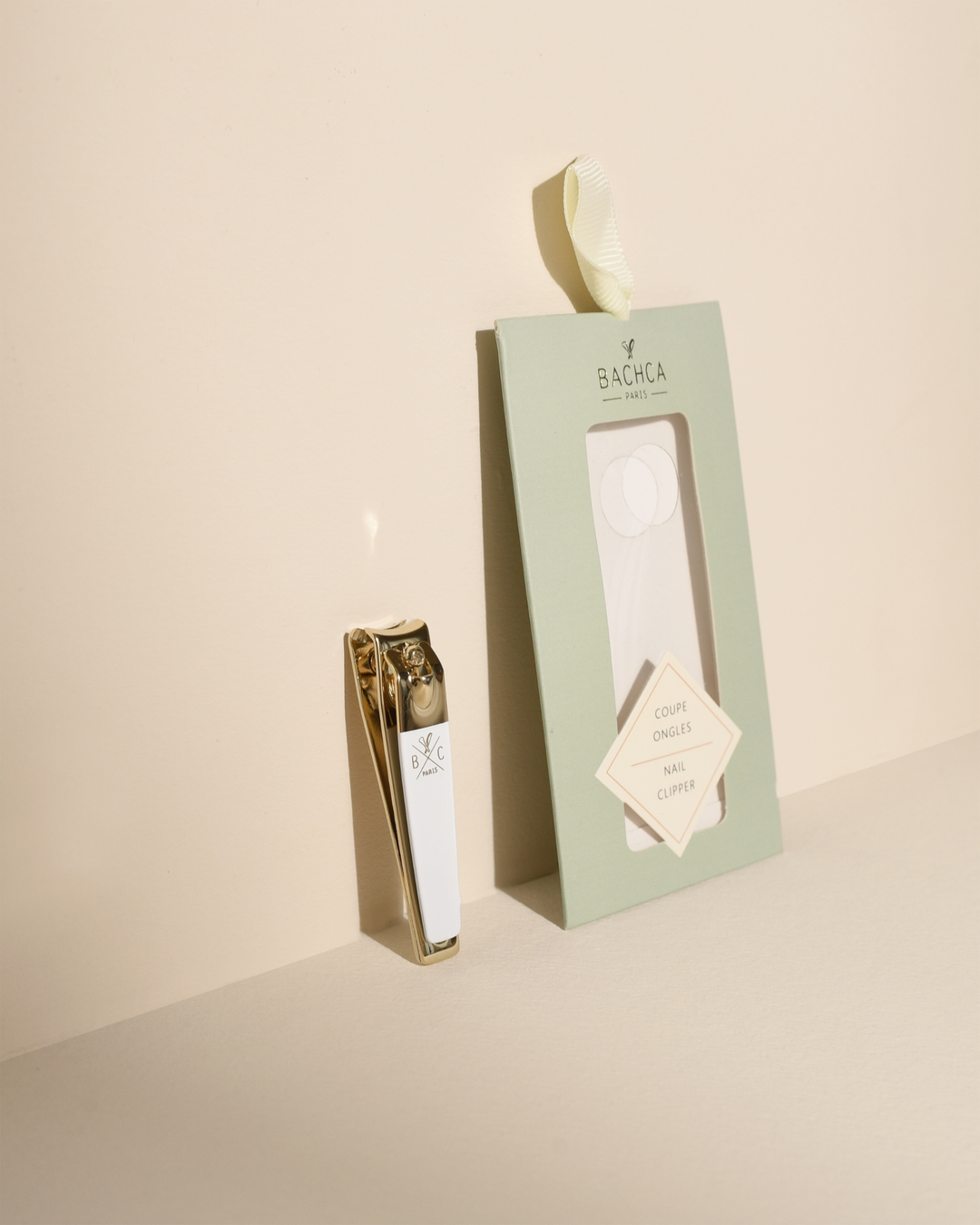 Gold and white nail clipper standing upright next to its empty sage green retail box on a warm beige background.