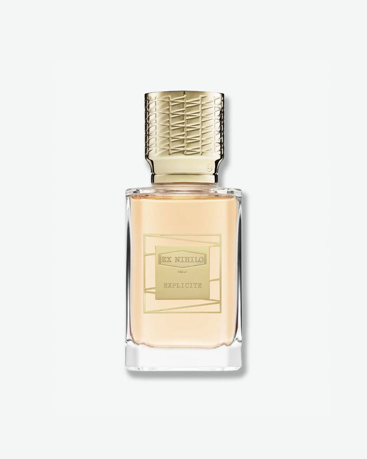 Ex Nihilo Explicite Eau de Parfum in a heavy clear glass bottle showcasing peach-tinted liquid, topped with an intricately engraved gold cap and featuring a matching gold geometric label, isolated on a light grey background.