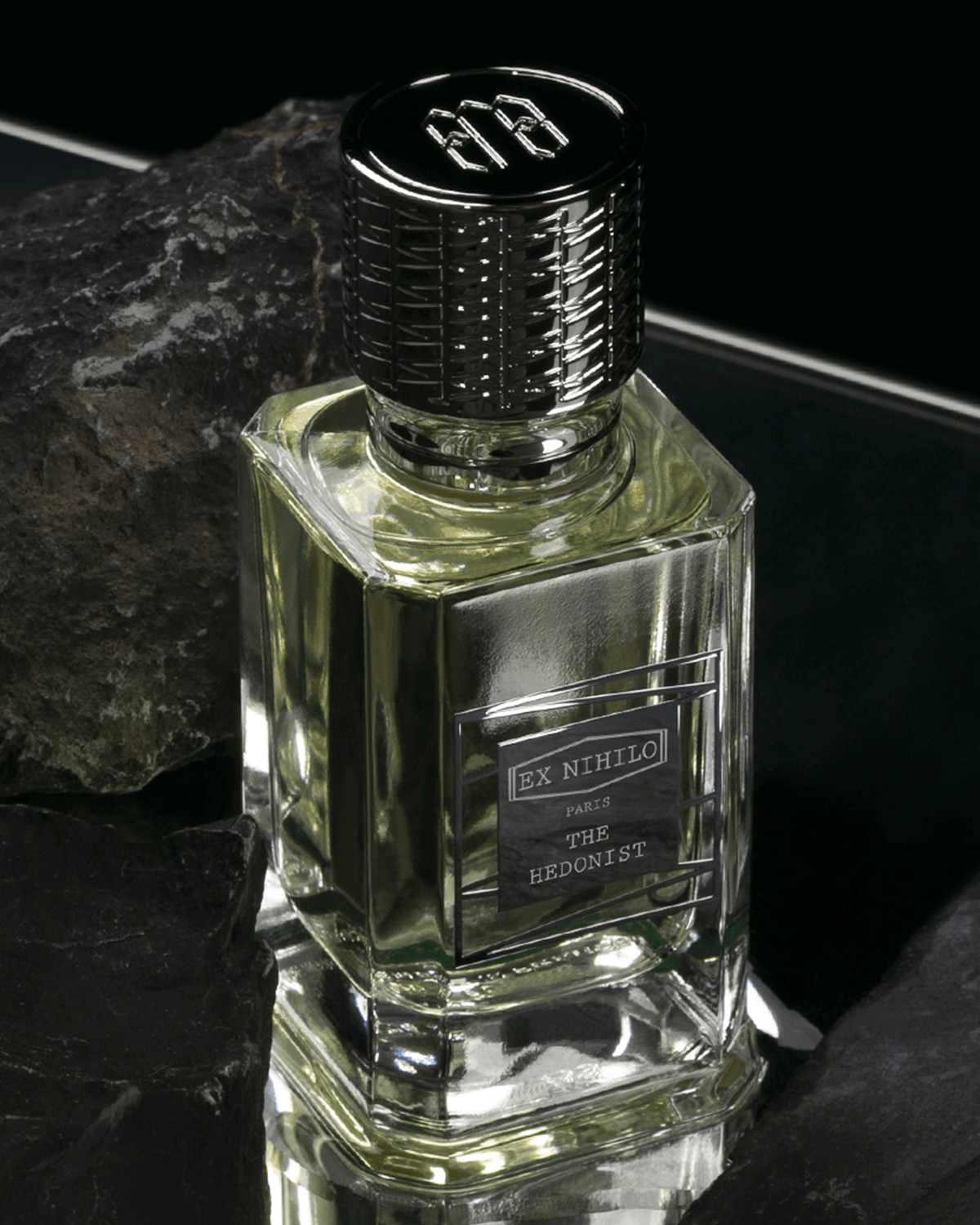 A clear glass perfume bottle with pale yellow liquid, a black ribbed cap, and a front label reading 'EX NTHIO PARIS THE LEONIST', photographed on a dark reflective surface with dark rocks around it.