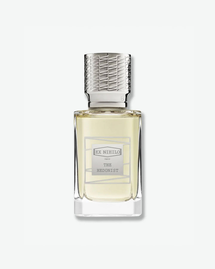 Ex Nihilo The Hedonist Eau de Parfum in a heavy clear glass bottle showcasing a pale chartreuse liquid, topped with an intricately engraved silver cap and a matching silver geometric label, isolated on a light grey background.