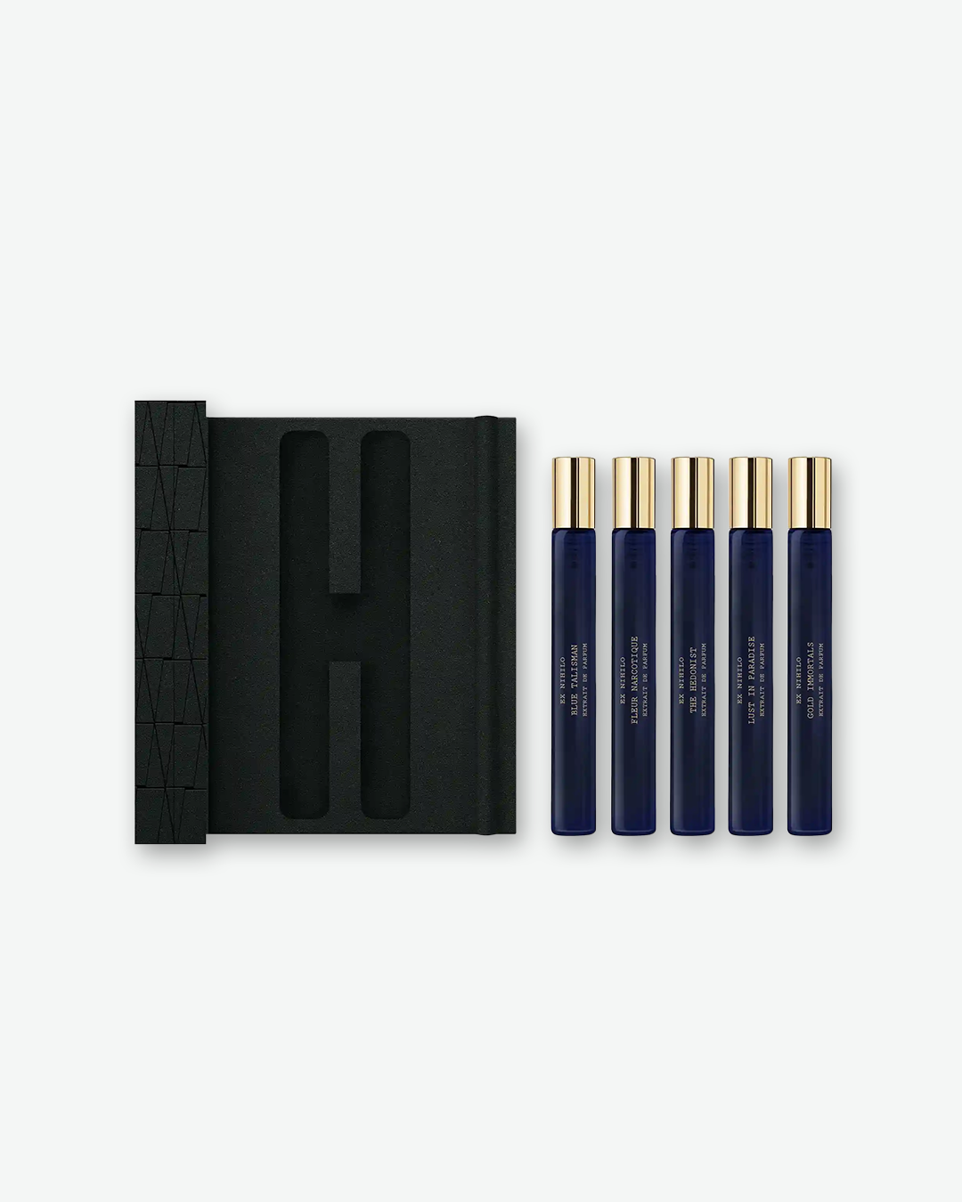 An overhead view of the open EX NIHILO travel set. On the left is the black foam-lined interior of the presentation box, showing molded slots for the fragrance vials. To the right, the five dark blue and gold-capped travel perfume vials (Blue Talisman, Fleur Narcotique, The Hedonist, Lust in Paradise, and Gold Immortals) are lined up vertically, displayed outside of the box against a clean, light grey background.