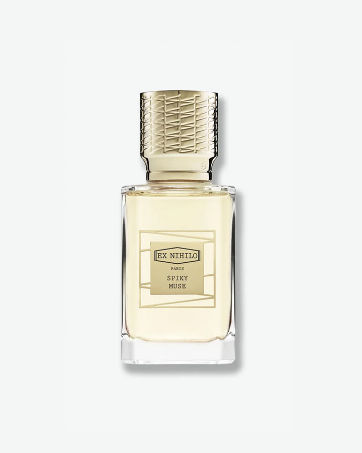 Ex Nihilo Spiky Muse Eau de Parfum in a heavy clear glass bottle showcasing a pale yellow liquid, topped with an intricately engraved gold cap and a matching gold geometric label, isolated on a light grey background.