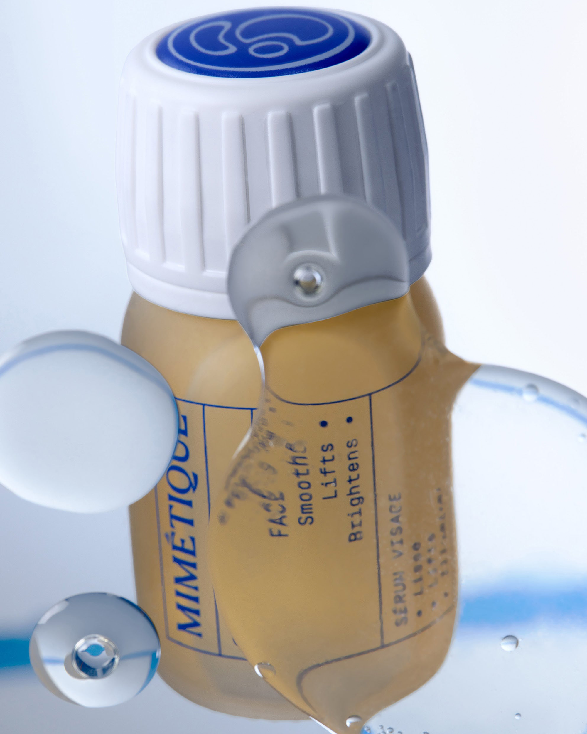 A close-up of a Mimétique facial serum bottle with a white ribbed cap and a clear dropper-style applicator resting on top. The bottle contains a pale yellow liquid and is photographed against a clean white background with reflective water droplets.