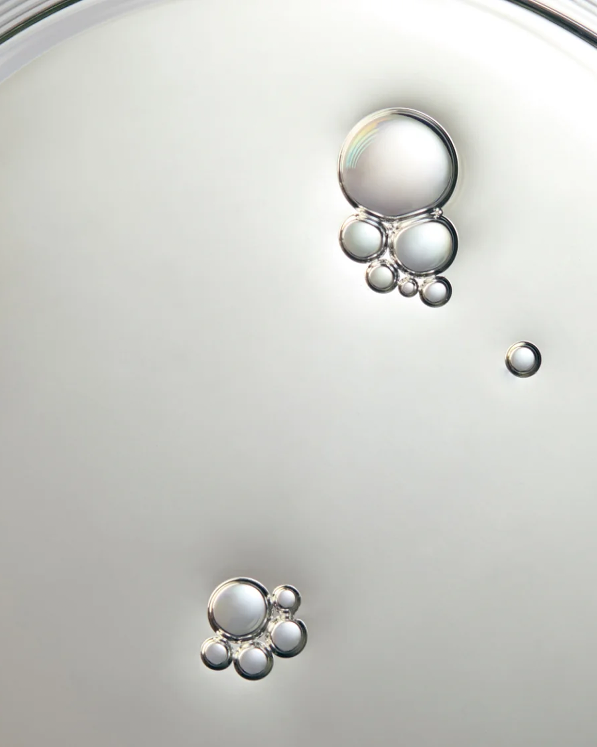 A close-up studio shot of a clear, glossy liquid droplet or oil drop resting on a smooth, light gray surface.