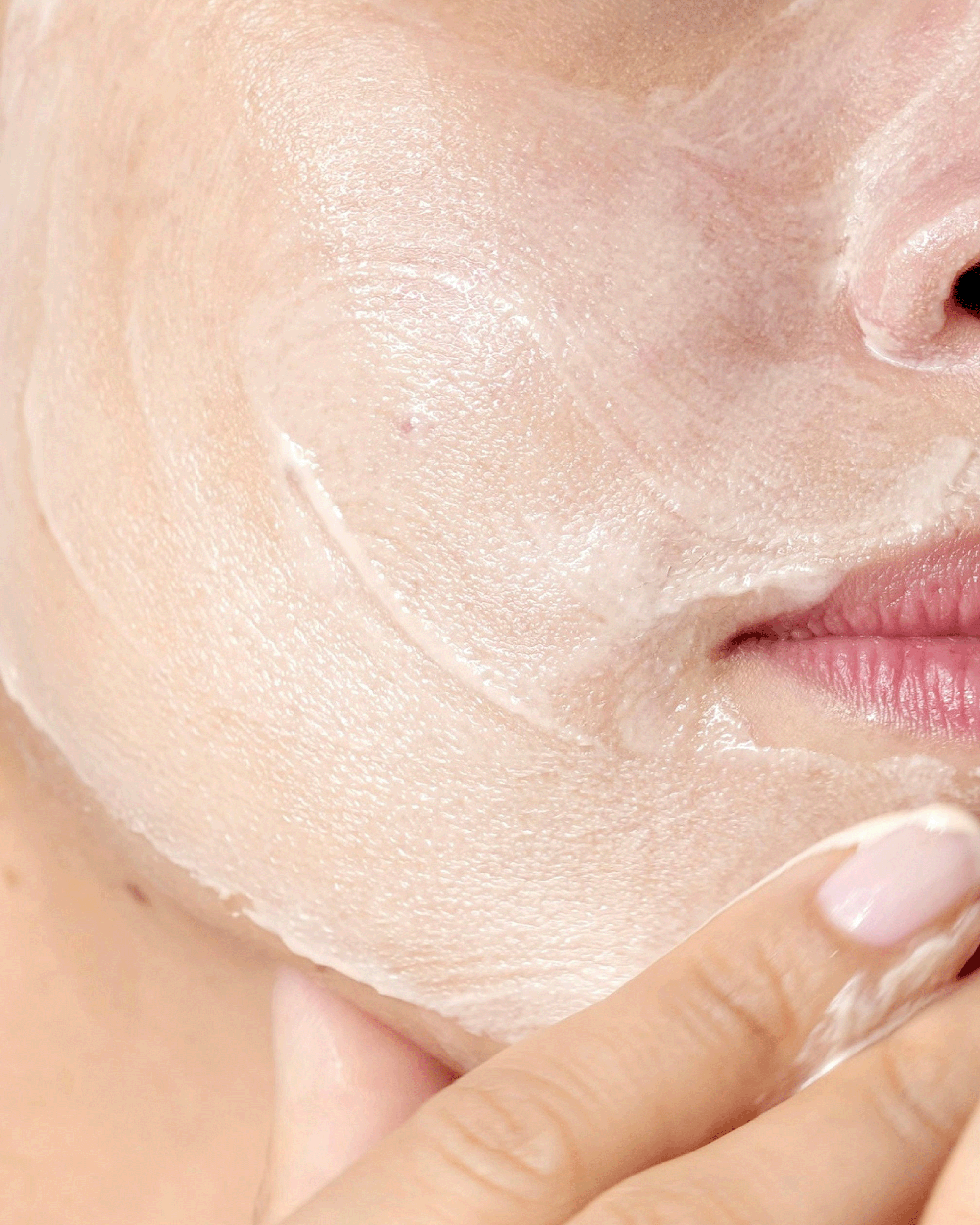 Close-up of a person's face and hand applying or smoothing a translucent, creamy skincare mask or gel on the cheek.