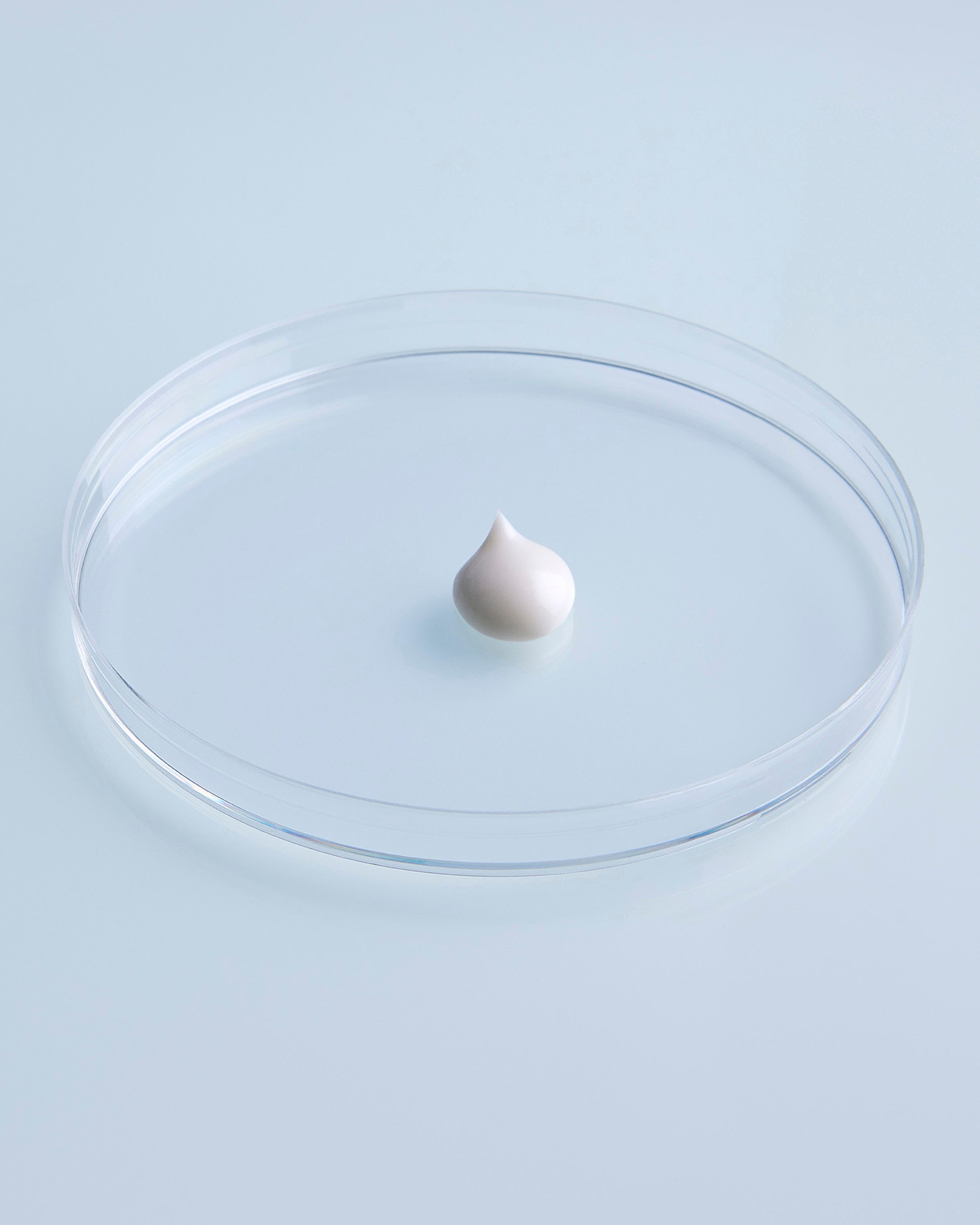 A small white cream dollop sits centered in a clear round petri dish against a pale blue background.