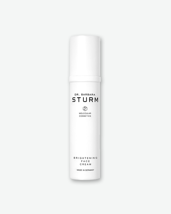 A product shot of a Dr. Barbara Sturm Brightening Face Cream bottle, centered against a light gray background. The sleek, slender cylindrical bottle is solid white with a matching opaque pump cap. The clean black text on the front reads "DR. BARBARA STURM", the molecular hexagon logo, "BRIGHTENING FACE CREAM", and "MADE IN GERMANY".