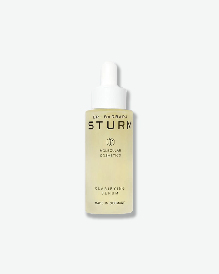 A product shot showing an open bottle of Dr. Barbara Sturm Clarifying Serum on a light gray background. On the right is the frosted glass bottle filled with a light yellow translucent liquid. Standing to its left is the unscrewed white dropper wand, revealing a clear glass pipette with a small droplet hanging at the tip.