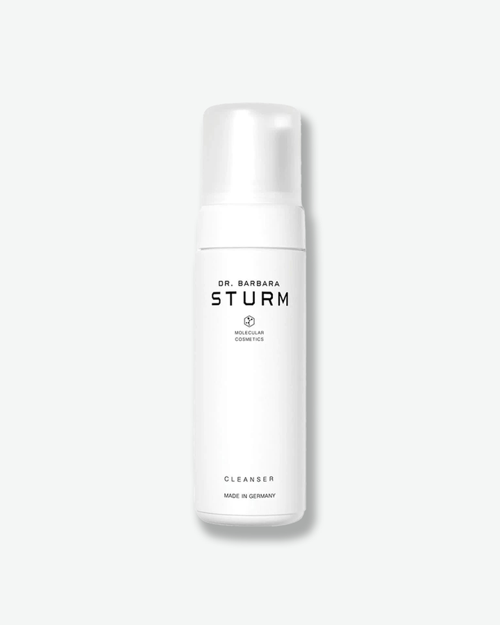 A product shot of a Dr. Barbara Sturm Cleanser bottle, centered against a plain light gray background. The tall cylindrical bottle is solid white and is topped with a matching white pump dispenser and a semi-translucent clear plastic cap. The front displays clean black text reading "DR. BARBARA STURM", the molecular hexagon logo, "CLEANSER", and "MADE IN GERMANY".
