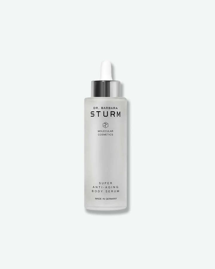 A product shot of a tall Dr. Barbara Sturm Super Anti-Aging Body Serum bottle, centered on a light gray background. The large cylindrical bottle is made of frosted glass, filled with a cloudy liquid, and topped with a metallic silver dropper cap and a white rubber bulb.