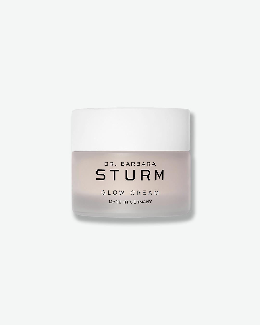 Front view of a Dr. Barbara Sturm Glow Cream jar against a plain gray background. The cosmetic container features a frosted translucent glass base through which a pale pinkish cream is visible, black branding text, and a clean white cap.