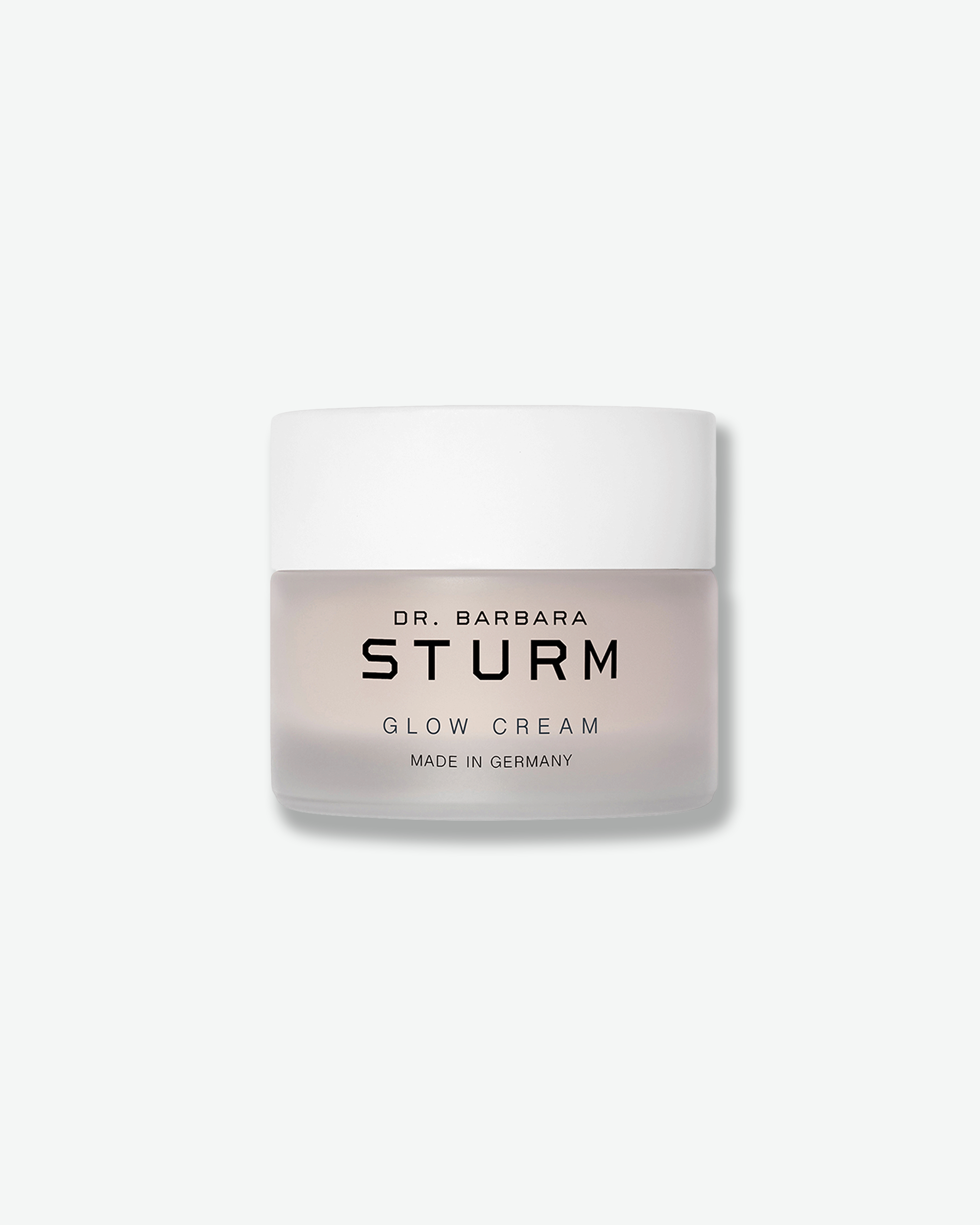 Front view of a Dr. Barbara Sturm Glow Cream jar against a plain gray background. The cosmetic container features a frosted translucent glass base through which a pale pinkish cream is visible, black branding text, and a clean white cap.