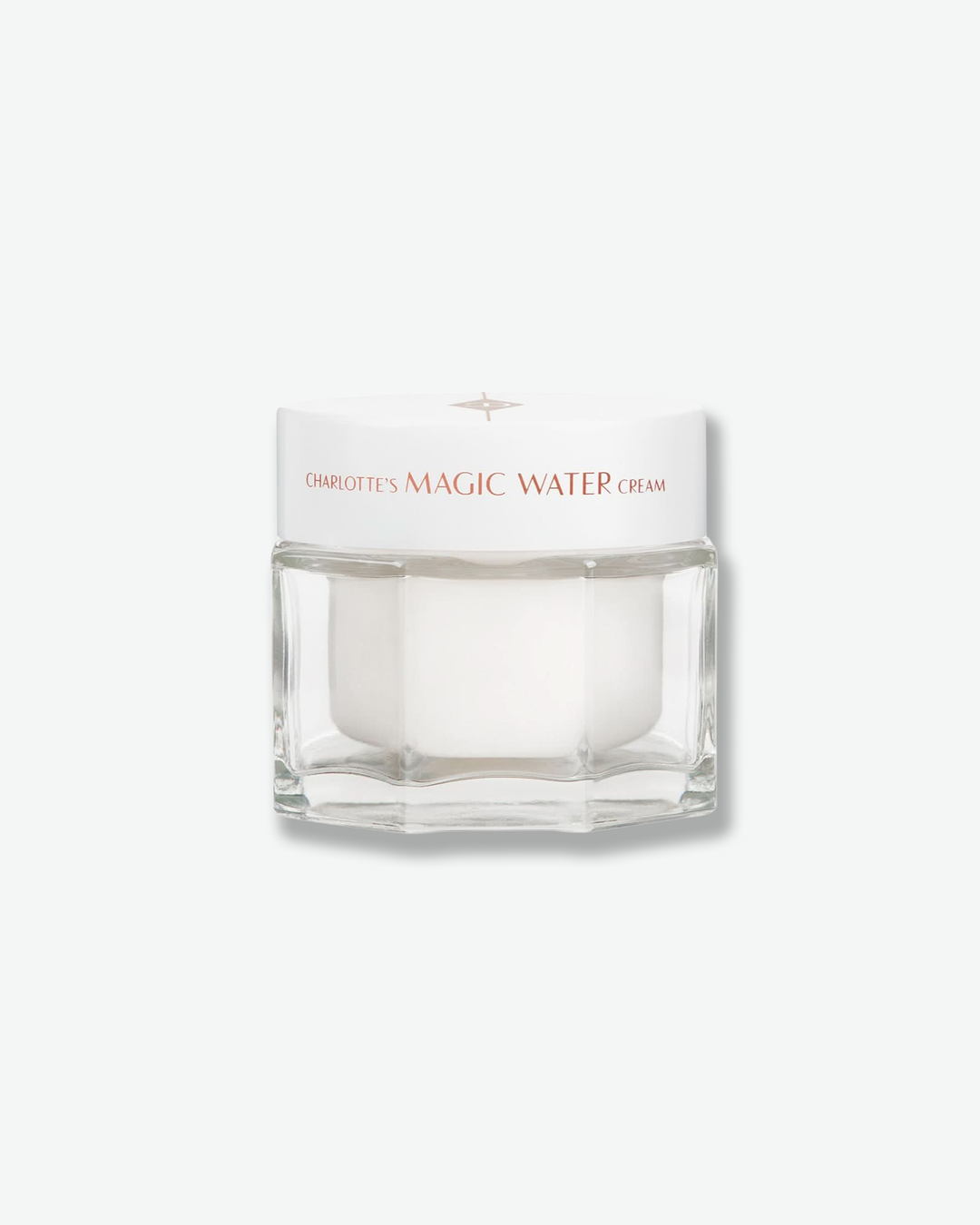 Front view of a Charlotte’s Magic Water Cream jar against a plain gray background. The product is housed in a clear, multi-faceted geometric glass jar with an opaque white inner pod, topped with a clean white screw-on lid featuring the product name in rose gold text.