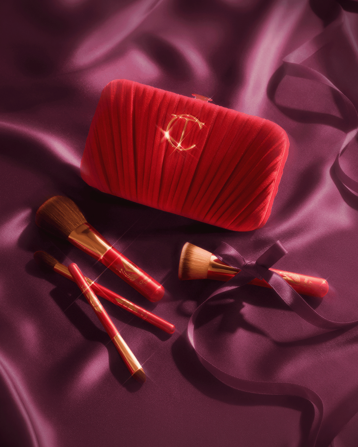 Charlotte Tilbury red velvet clutch and four mini makeup brushes arranged on deep berry satin.