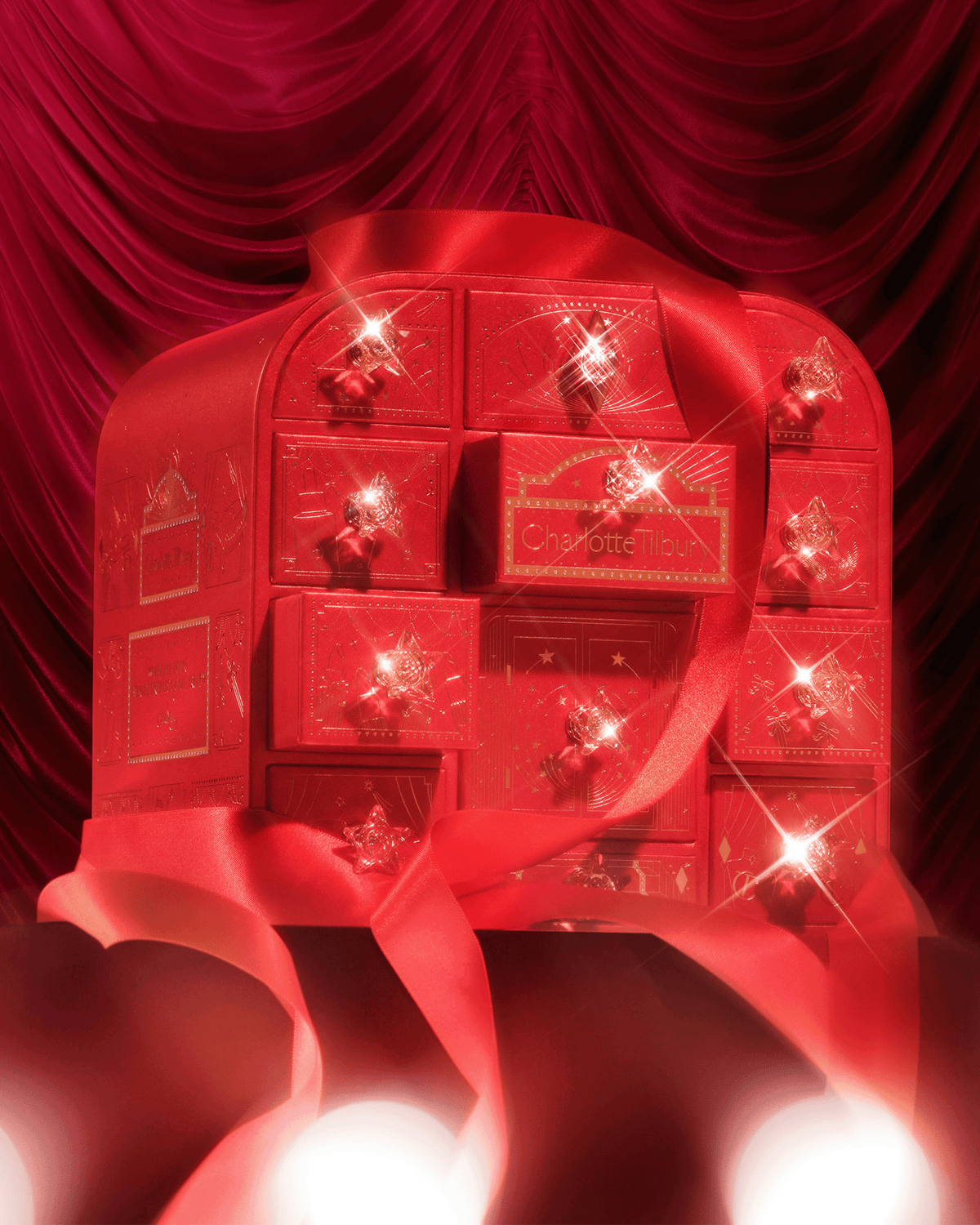 Charlotte Tilbury Beauty Treasure Chest of Love styled with red satin ribbon and sparkling holiday details against a dramatic red curtain backdrop.