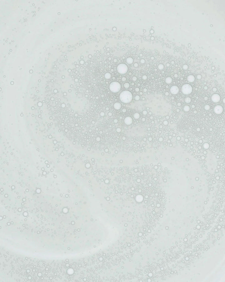 An abstract close-up of a milky skincare formula featuring hundreds of suspended microbubbles. The lightweight emulsion is shown in a swirling pattern against a pale background, highlighting its smooth texture and blend of oil-and-water components.