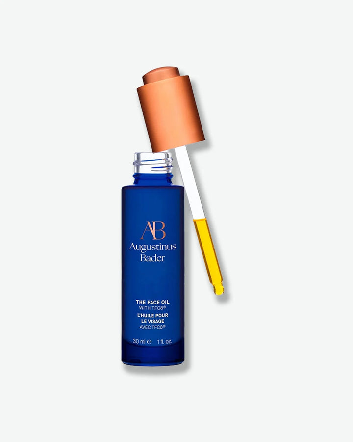 An open bottle of Augustinus Bader The Face Oil in a deep blue glass container, with the copper-toned dropper pipette resting outside, showing a drop of golden oil.