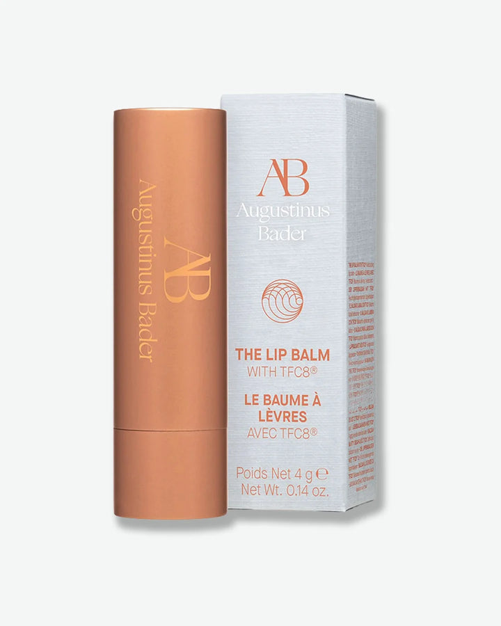 Bronze colored Augustinus Bader lip balm stick with cap on stood next to its light grey packaging box on a lighter grey background