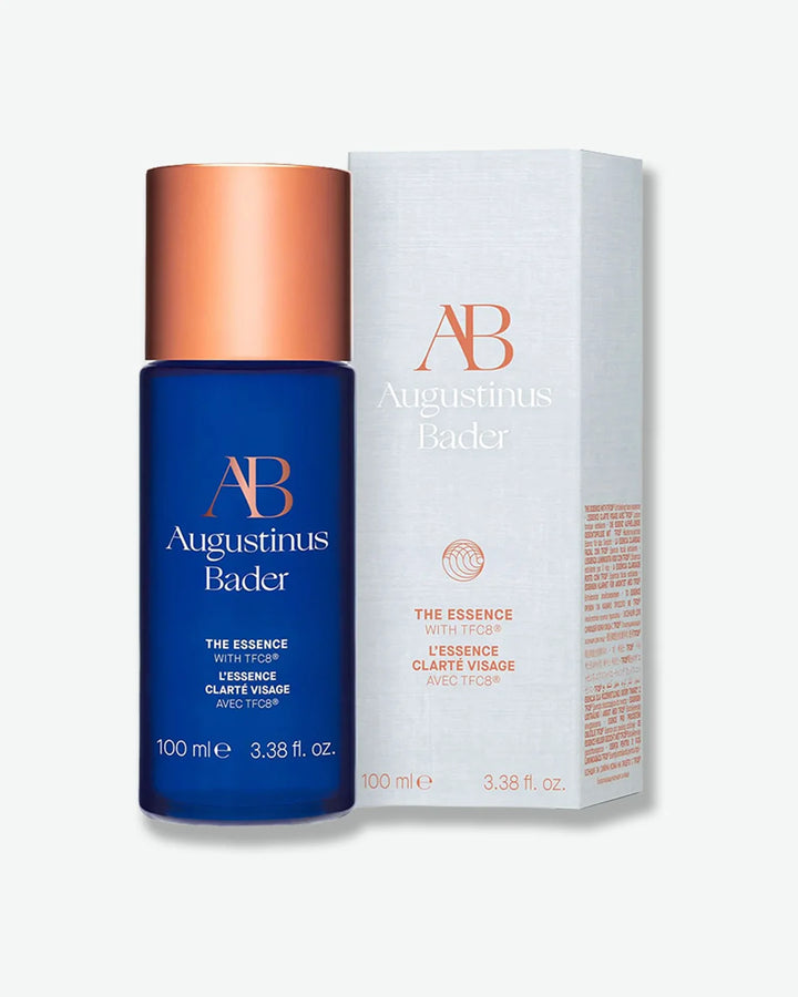 Blue Augustinus Bader The Essence bottle with a copper cap, standing next to its light gray product packaging box on a white background.