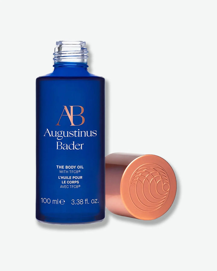 Blue Augustinus Bader The Body oil bottle with a copper screw-top cap off next to it, on a light gray background.