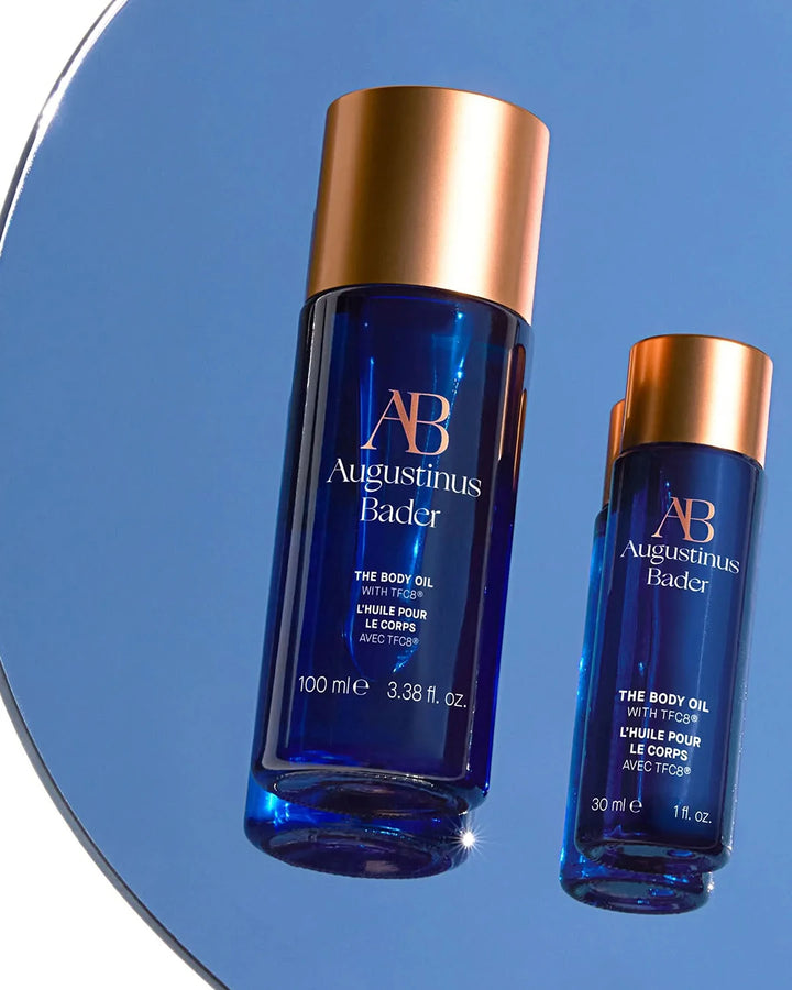 Two blue bottles of Augustinus Bader body oil with gold caps on a reflective surface.