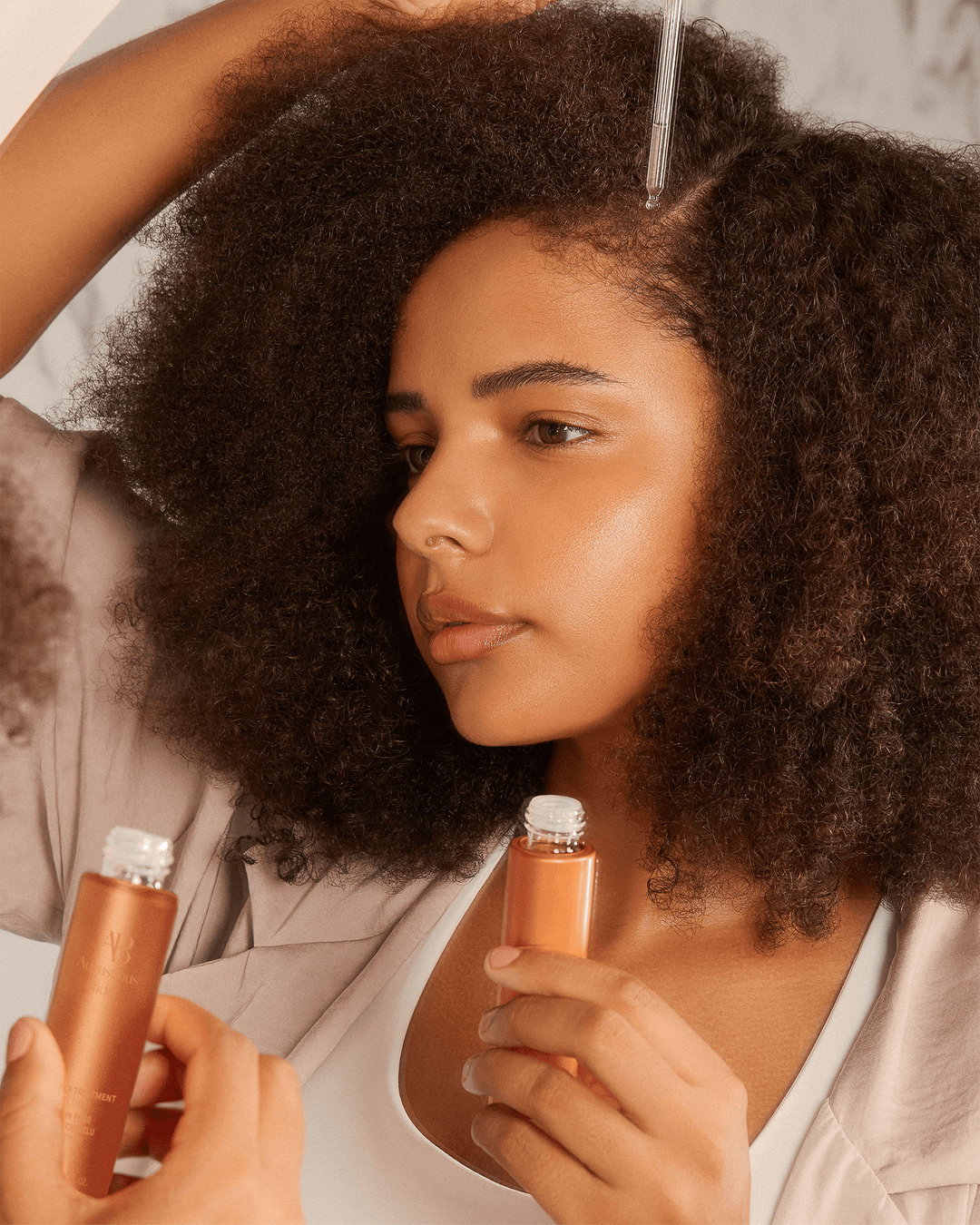 A person with curly hair holding a copper-colored bottle while using a glass dropper to apply the clear treatment directly to their scalp.