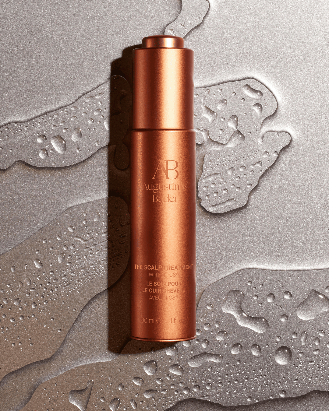 An Augustinus Bader copper-colored scalp treatment bottle lying flat on a shimmering, wet metallic silver surface covered in water droplets.