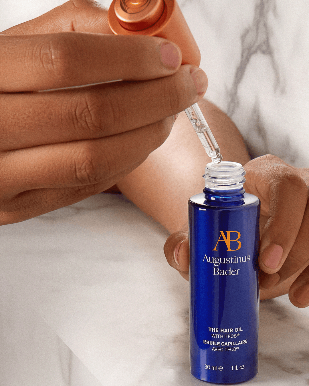 A person's hands holding a blue Augustinus Bader hair oil bottle while lifting the dropper out, showcasing the clear product.