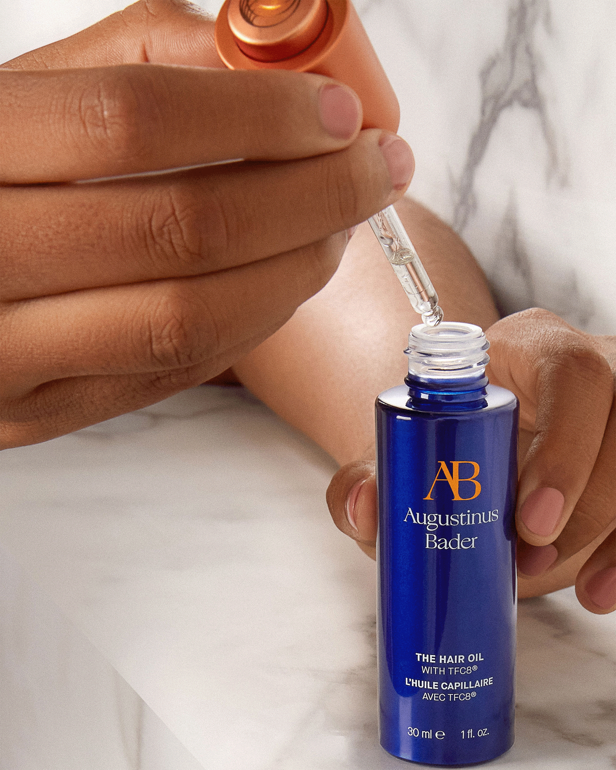 A person's hands holding a blue Augustinus Bader hair oil bottle while lifting the dropper out, showcasing the clear product.