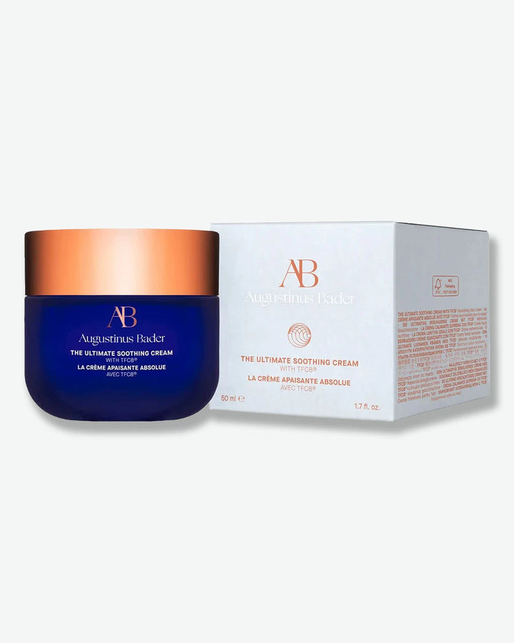 Augustinus Bader The Ultimate Soothing Cream jar with blue body and bronze lid stood next to its light grey packaging box with bronze text on a white background