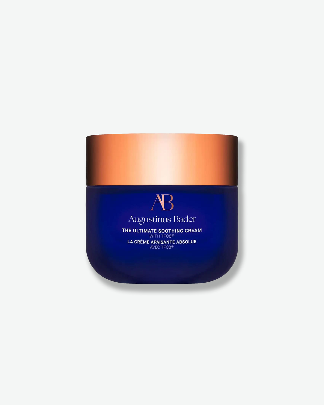 Front view of an Augustinus Bader "The Ultimate Soothing Cream" jar against a plain gray background. The design features a smooth, deep royal blue base with rounded bottom corners, showcasing copper-gold logo accents and white text branding, topped with a wide, metallic copper-gold cap.