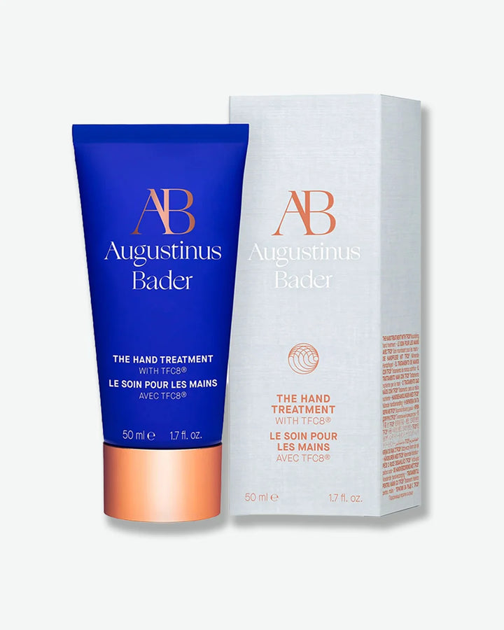 Blue Augustinus Bader The Hand Treatment tube with a copper cap, stood next to its light gray packaging box, against a lighter grey background.