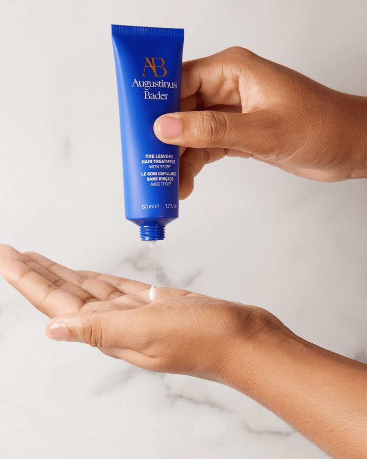 A person holding the blue Augustinus Bader leave-in hair treatment tube and squeezing a small dab of white cream into their open palm.