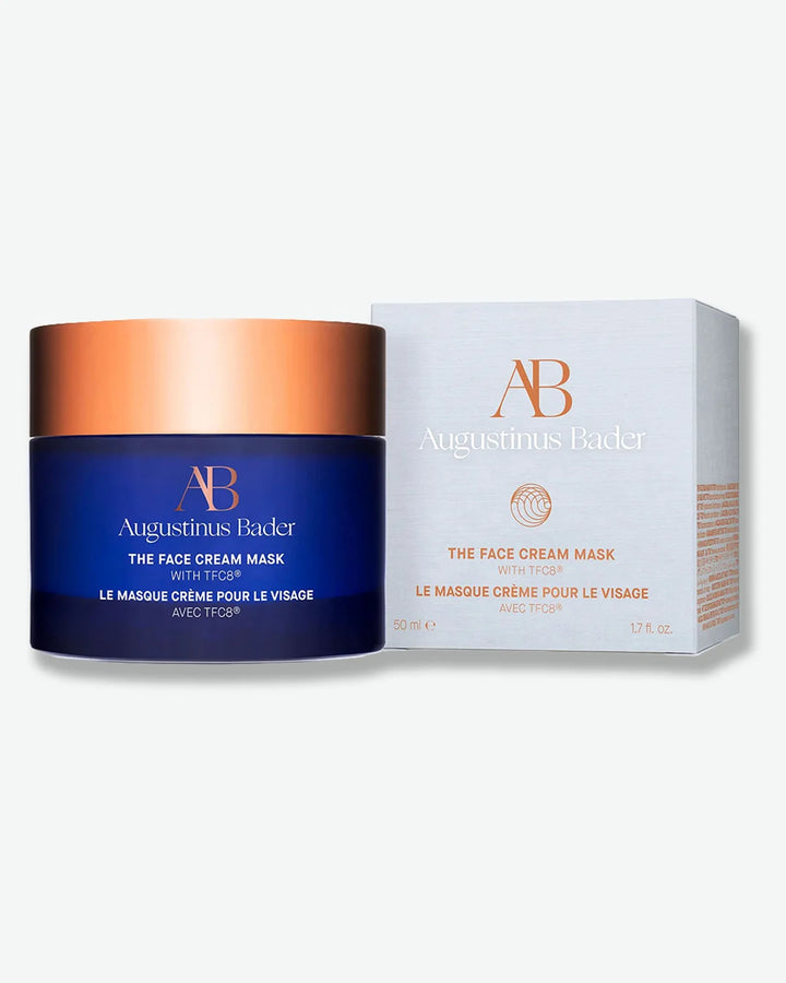 Augustinus Bader the face cream mask jar with blue body and bronze lid, stood next to its white backgrounds light grey packaging box, on a light grey background.