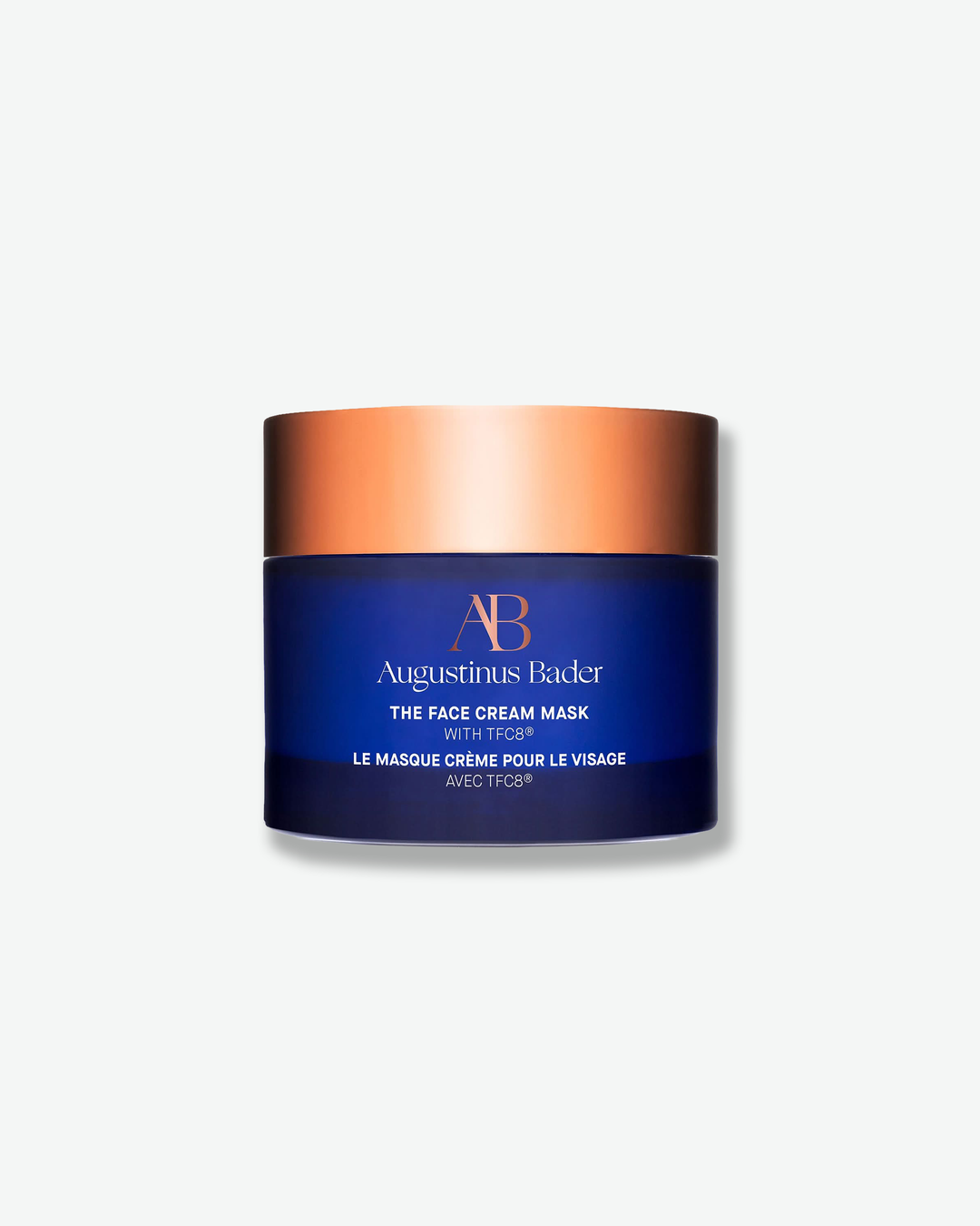 Front view of an Augustinus Bader "The Face Cream Mask" jar centered against a plain gray background. The cylindrical container consists of a dark royal blue body with minimalist copper and white branding, topped with a flat, metallic copper-gold screw lid.