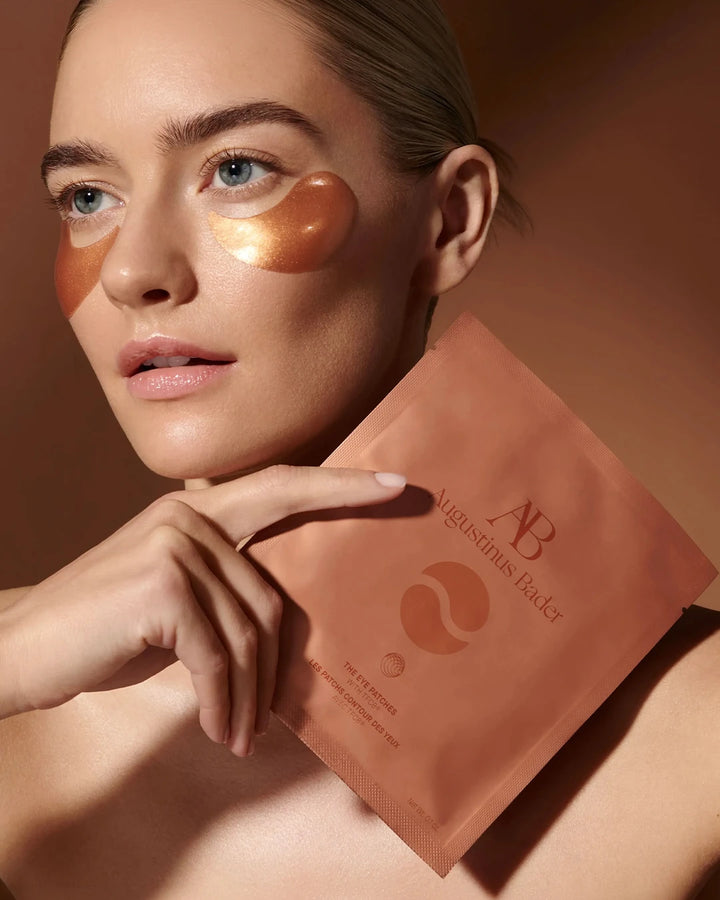 Woman applying gold eye patches with a product packaging on a warm-toned background