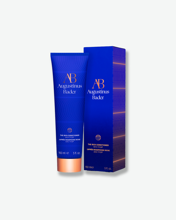 An Augustinus Bader blue conditioner tube standing next to its matching blue outer cardboard packaging box.