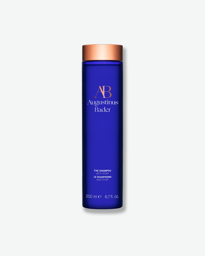 A tall, cylindrical dark blue bottle of Augustinus Bader The Shampoo with a metallic copper cap, centered on a light gray background.