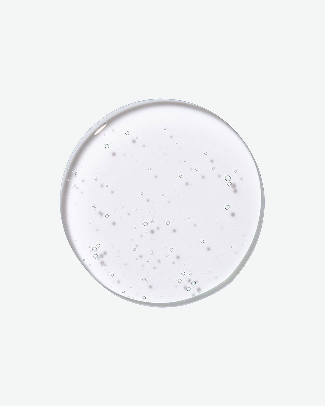 A top-down close-up view of a clear, circular droplet of shampoo filled with tiny air bubbles against a solid light gray background.