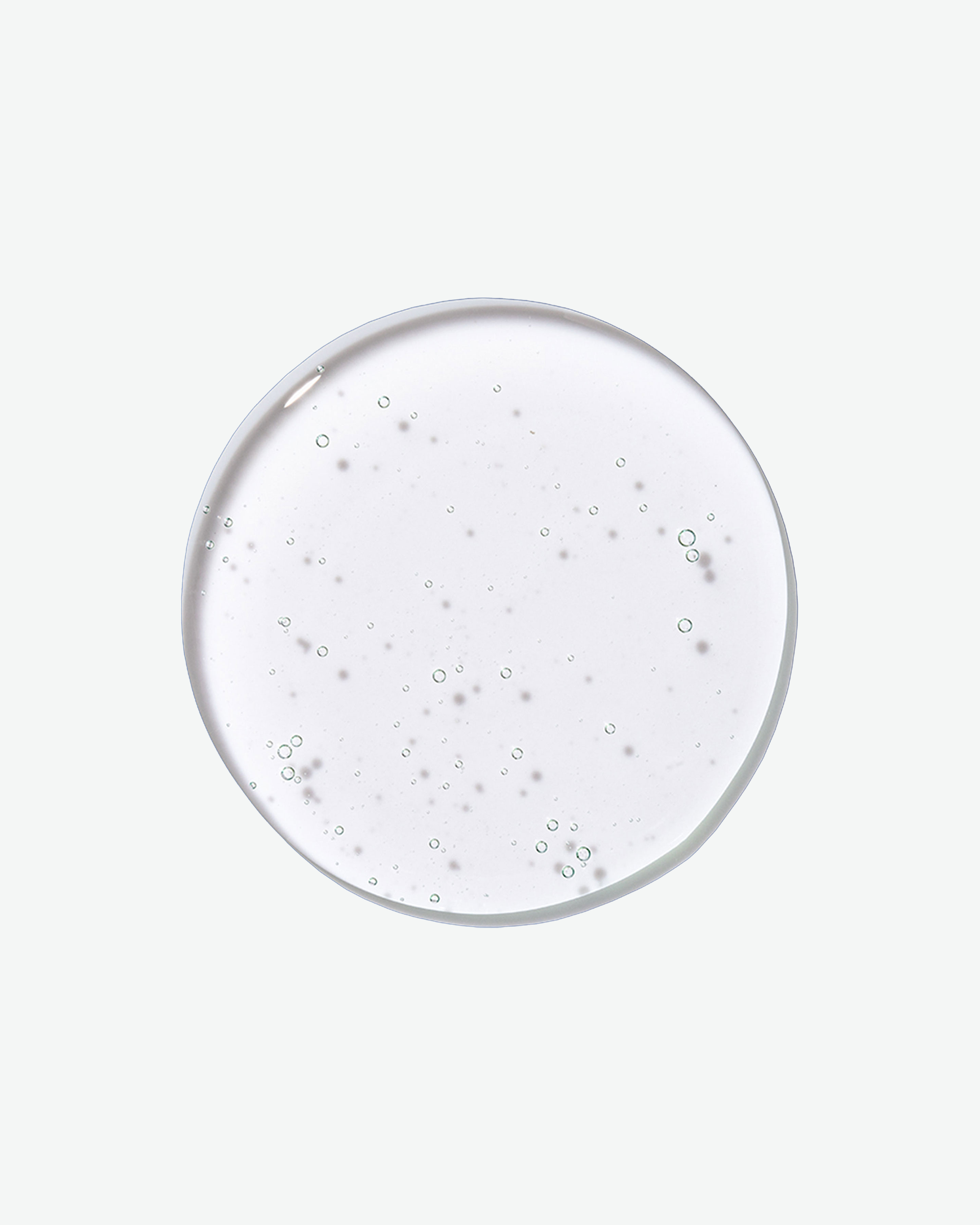 A top-down close-up view of a clear, circular droplet of shampoo filled with tiny air bubbles against a solid light gray background.
