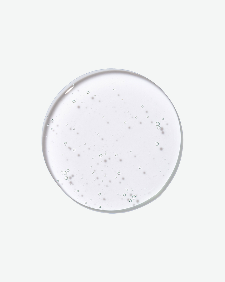 A top-down close-up view of a clear, circular droplet of shampoo filled with tiny air bubbles against a solid light gray background.