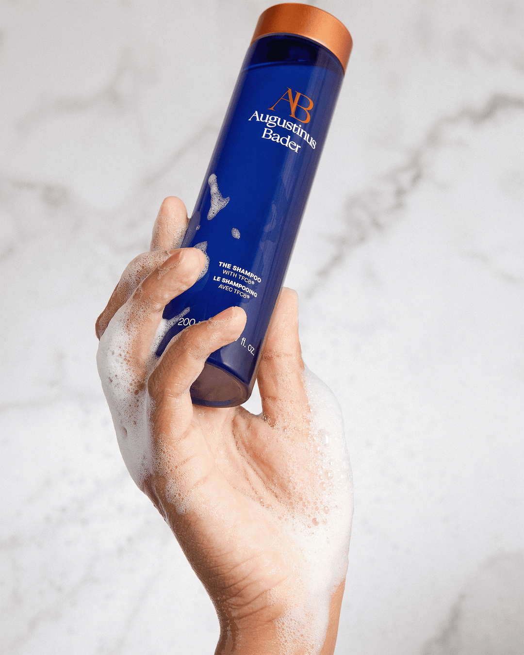 A soapy, foam-covered hand holding the blue Augustinus Bader shampoo bottle against a marbled gray background.