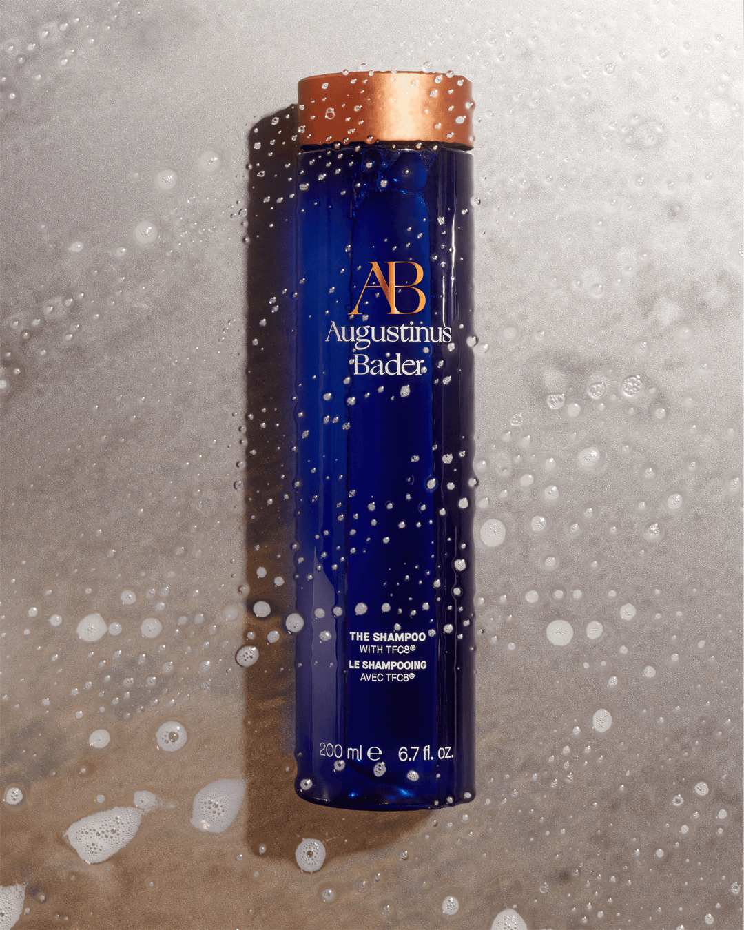 An Augustinus Bader blue shampoo bottle lying flat on a textured gray stone surface covered in water droplets and soapy white lather.