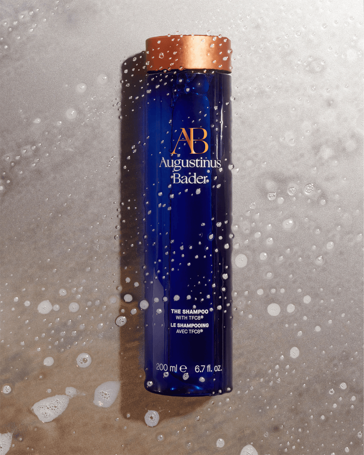 An Augustinus Bader blue shampoo bottle lying flat on a textured gray stone surface covered in water droplets and soapy white lather.