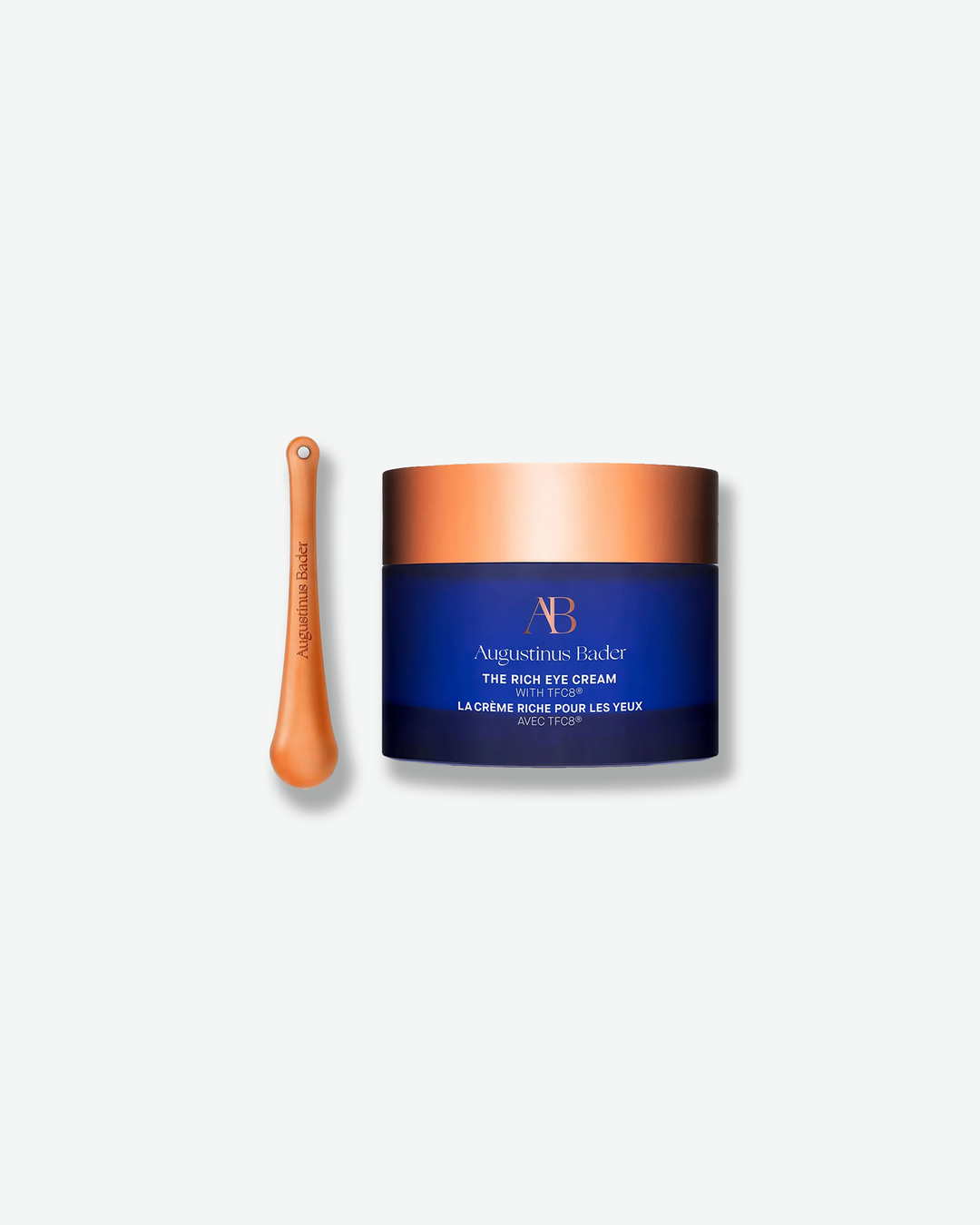 Product shot featuring an Augustinus Bader "The Rich Eye Cream" jar standing next to its application tool against a plain gray background. The royal blue cylindrical jar with a wide, metallic copper-gold lid is positioned beside a slender, matching copper-gold metallic massage spoon embossed with the brand name.