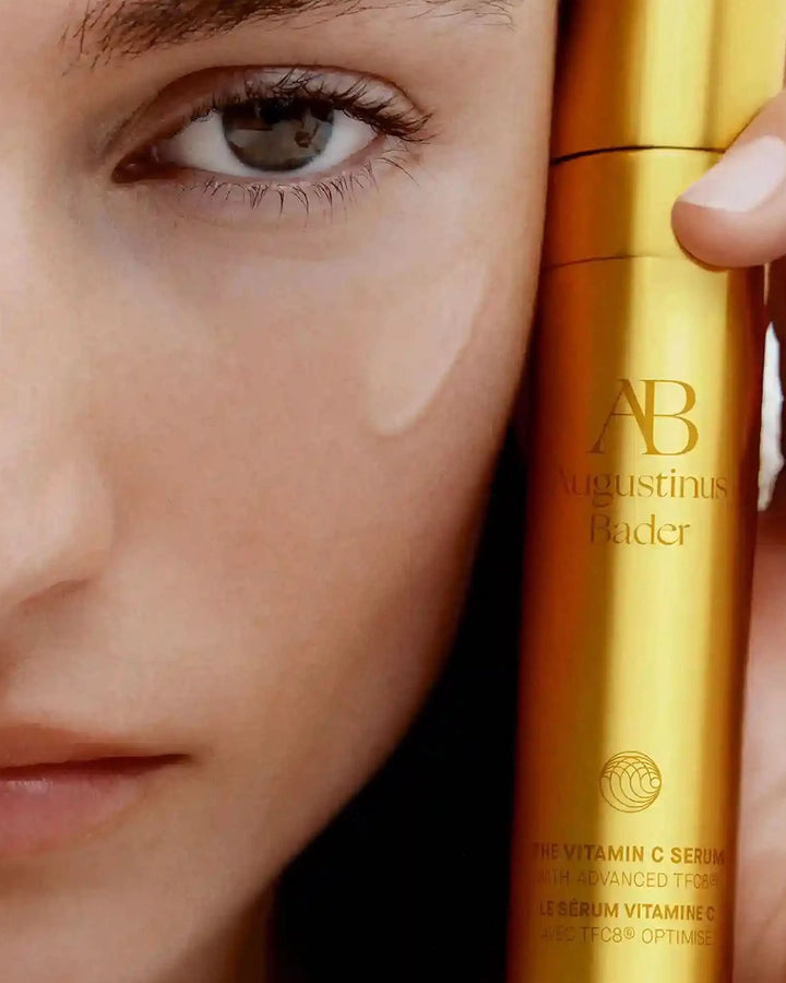 Close-up of a woman's face with a gold skincare bottle labeled 'Augustinus Bader' held close to her eye.