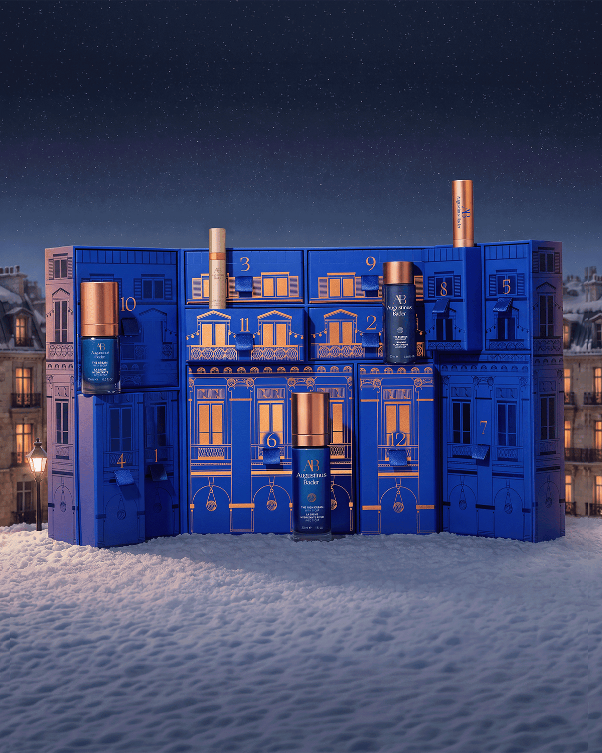 Augustinus Bader The 12 Days of Bader advent calendar displayed in a snowy nighttime city scene with select skincare products featured in front.