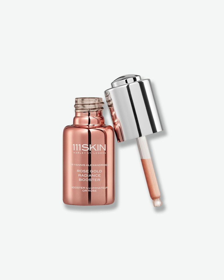 Product shot of an open 111SKIN Rose Gold Radiance Booster bottle against a plain gray background. The small, cylindrical glass bottle has a highly reflective, metallic rose gold finish with white branding. Propped up next to the open neck is its detached glass dropper cap, featuring a high-shine silver metallic collar and a tint of peach-colored booster liquid visible inside the pipette.