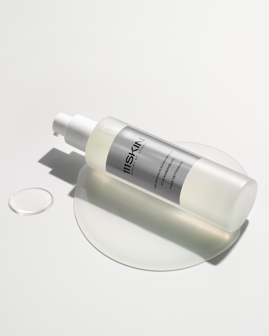 A 111SKIN cleansing oil bottle resting on a white surface amidst a puddle and a droplet of clear oil.